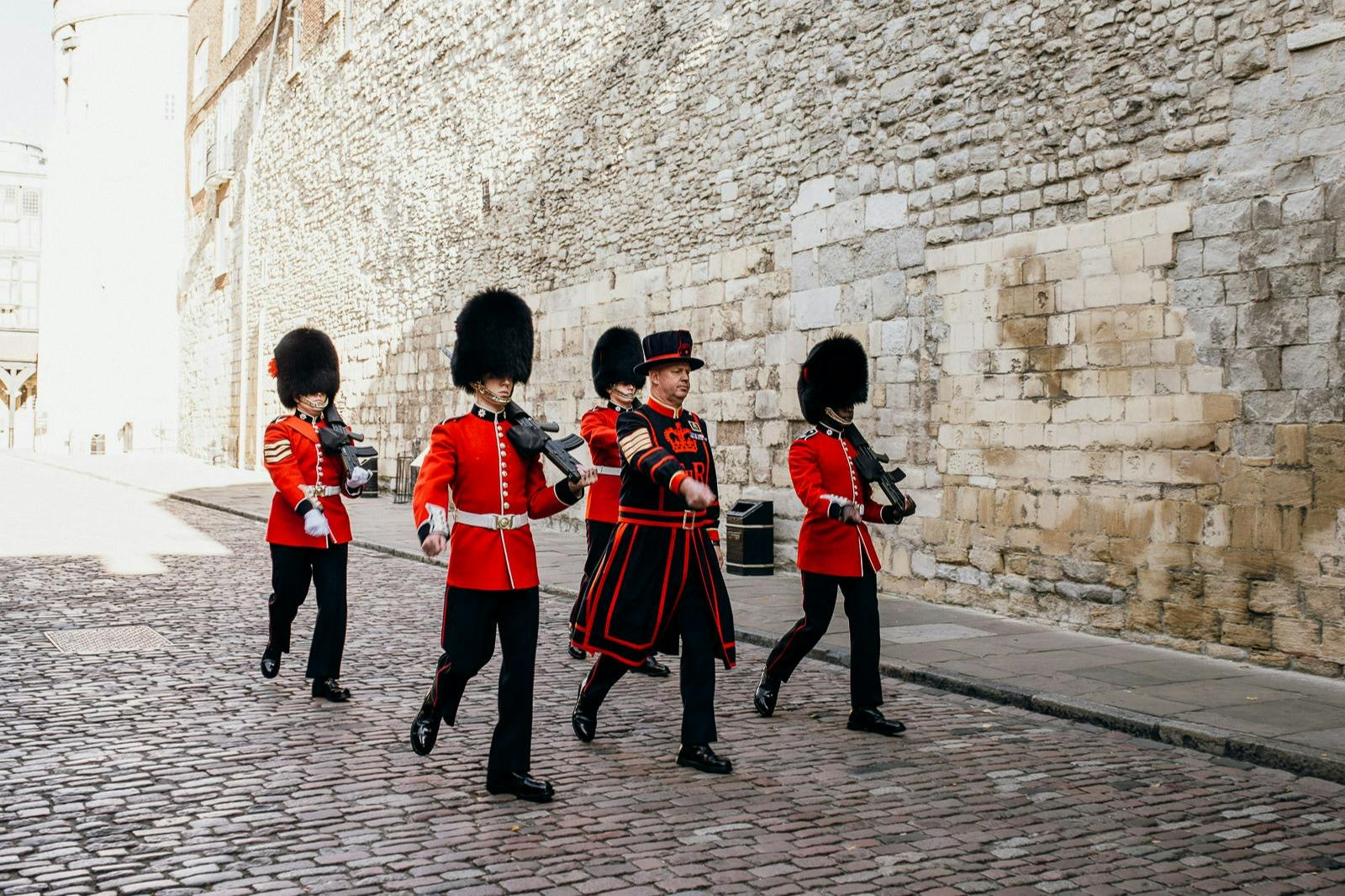 Uniformed guards, with bearskin hats and red coats, march on a cobblestone street beside a stone wall.