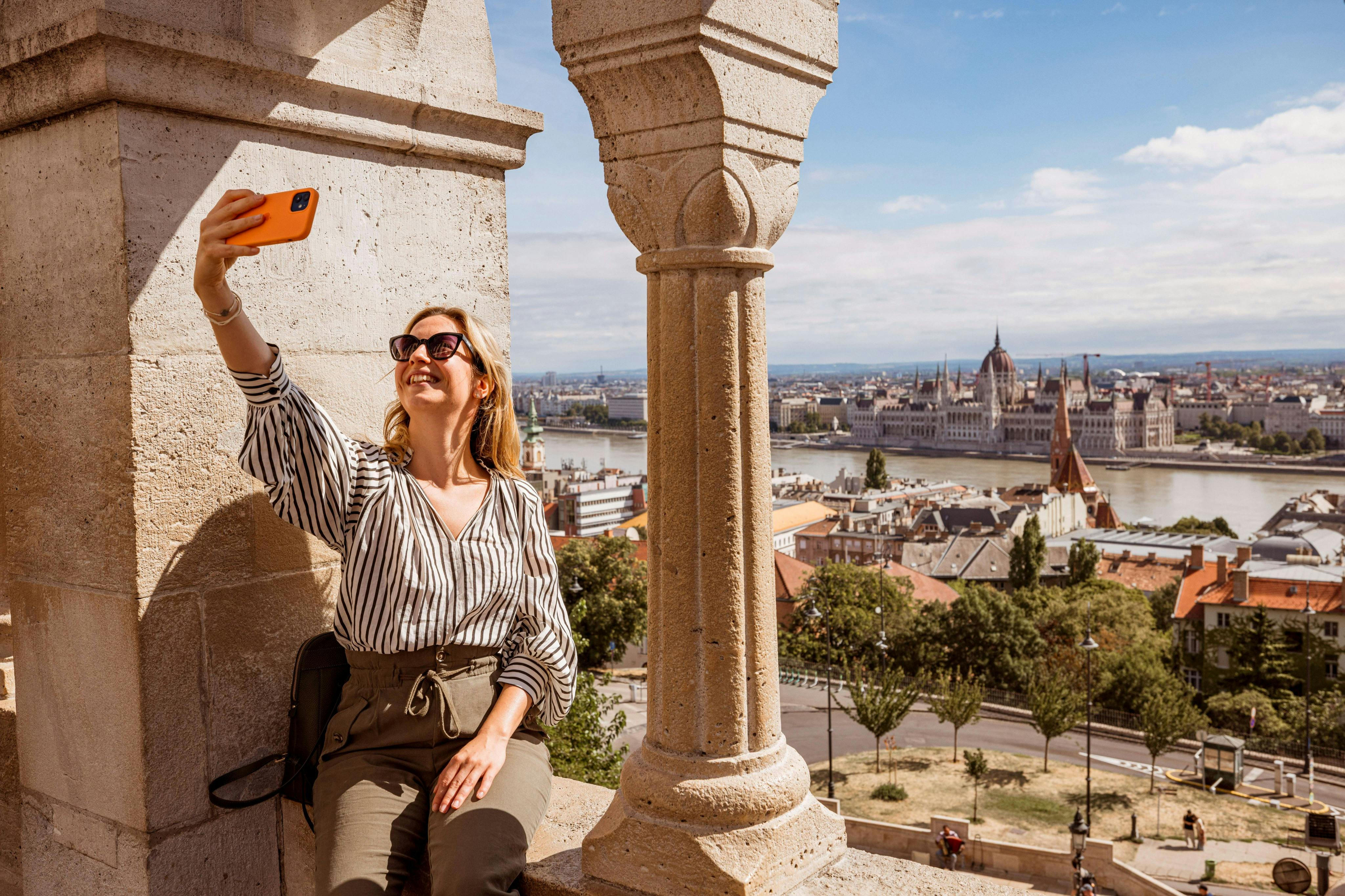 A woman wearing sunglasses takes a selfie with a smartphone while sitting near a stone column overlooking a cityscape.