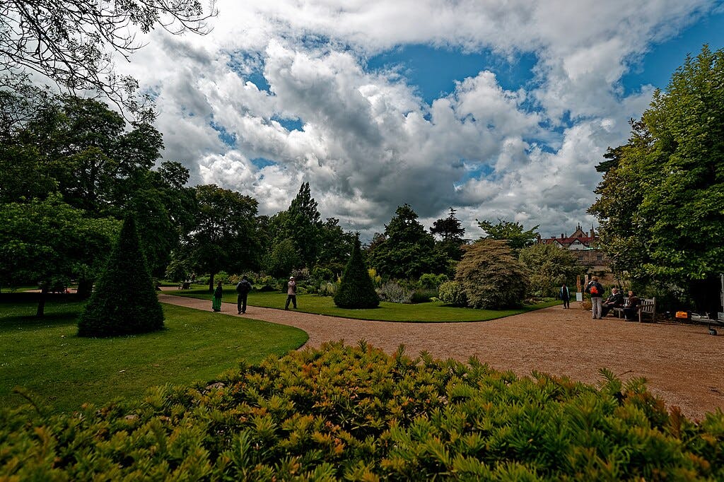 People walking and sitting on benches in a garden with pathways, trees, and trimmed bushes under a partly cloudy sky.