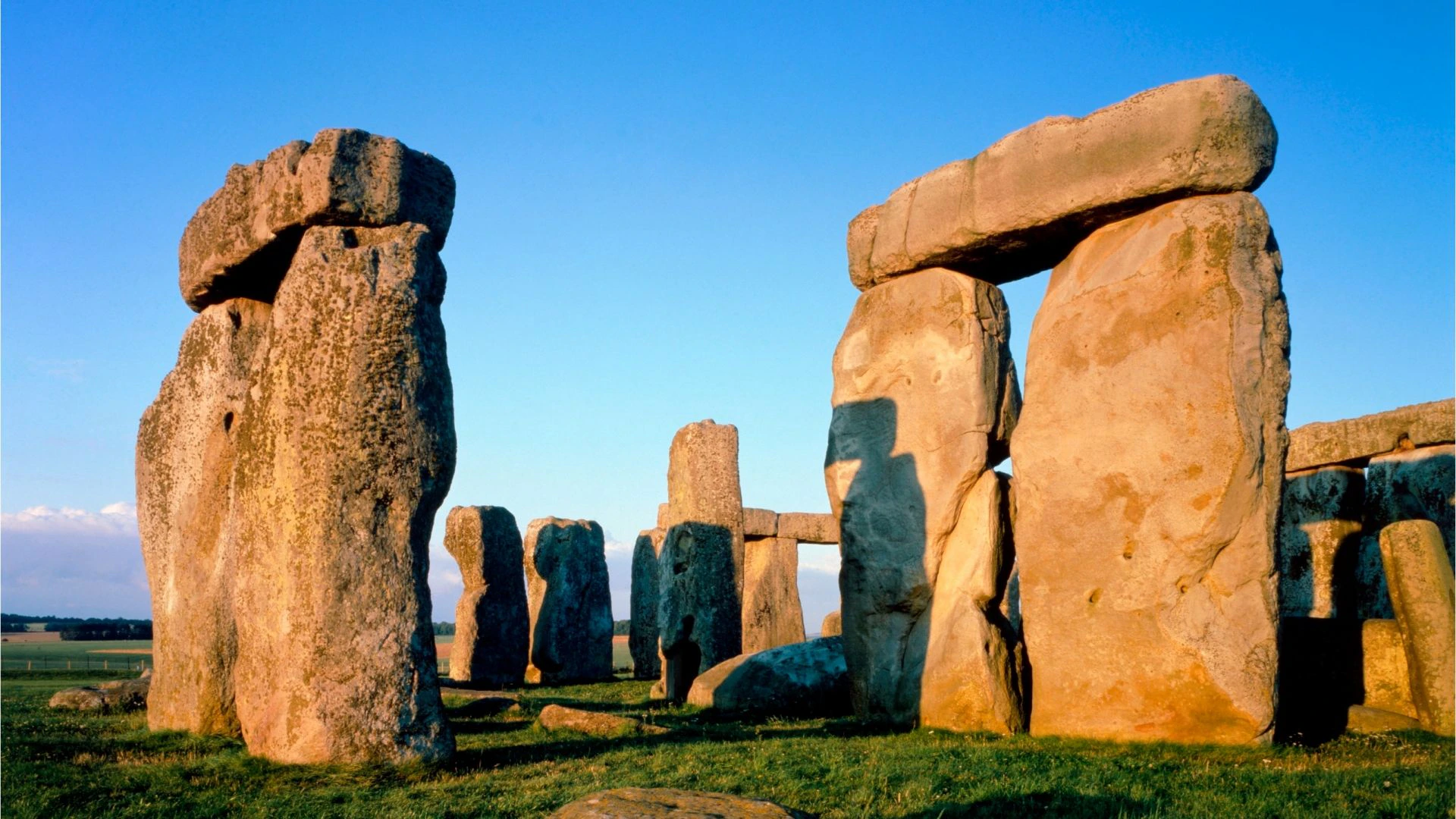 Large stone monoliths forming a circle on grass, under a clear blue sky, likely part of an ancient structure.