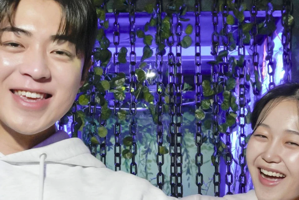 Two people smiling on a rope bridge in a setting with greenery and purple lighting.