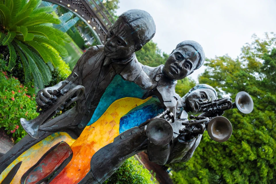 Three bronze statues of musicians playing instruments with colorful abstract designs. Greenery and a bridge in the background.