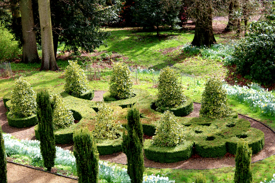 Well-maintained garden with intricately shaped hedges, small bushes, and a surrounding grassy area with large trees in the background.