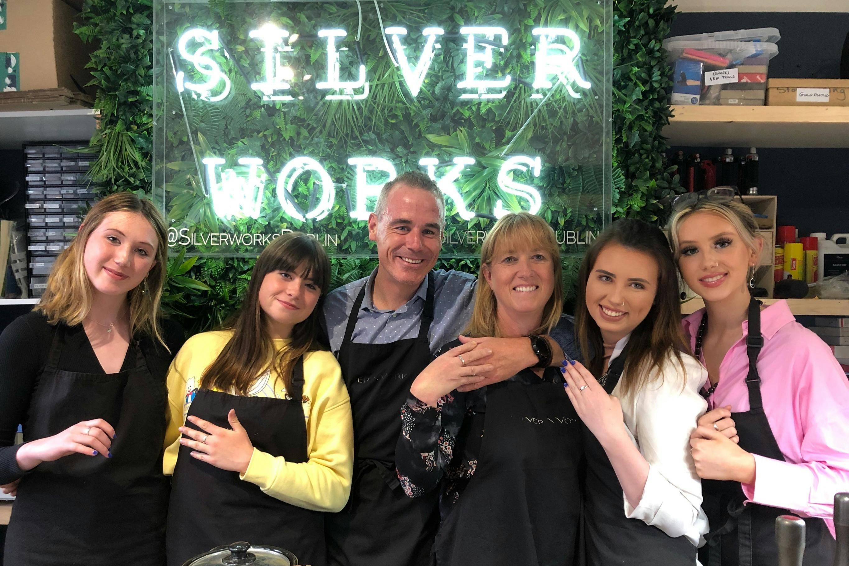 Five people in aprons smile and embrace in front of a greenery backdrop with the illuminated text "Silver Works."