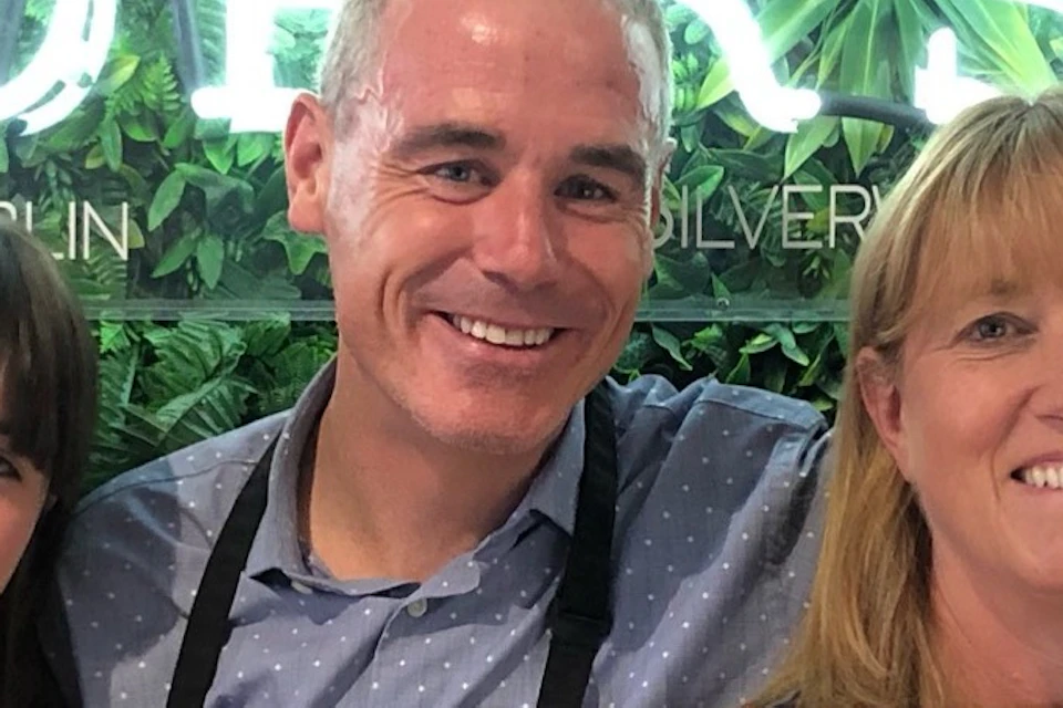 Five people in aprons smile and embrace in front of a greenery backdrop with the illuminated text "Silver Works."