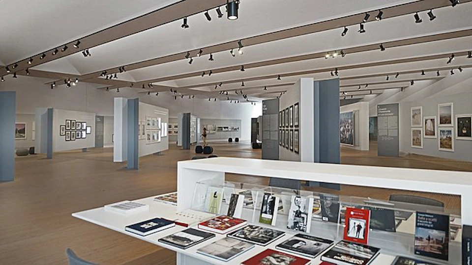 Modern gallery interior with art books on a white display table, framed photographs on walls, and ceiling spotlights.