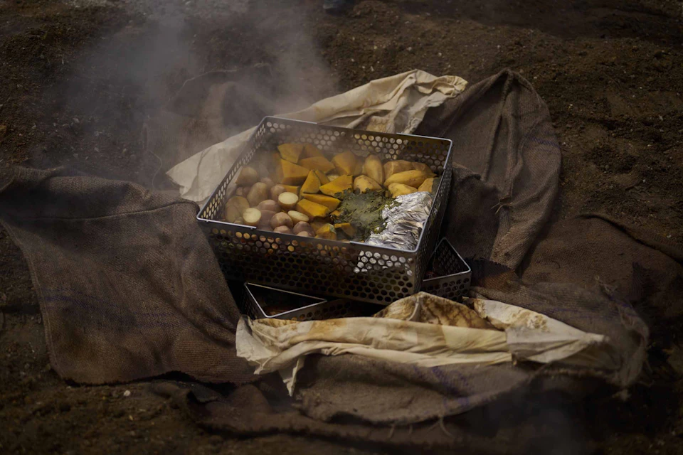 A metal container with cooked vegetables, foil, and herbs, surrounded by cloth, emitting steam, placed on the ground.