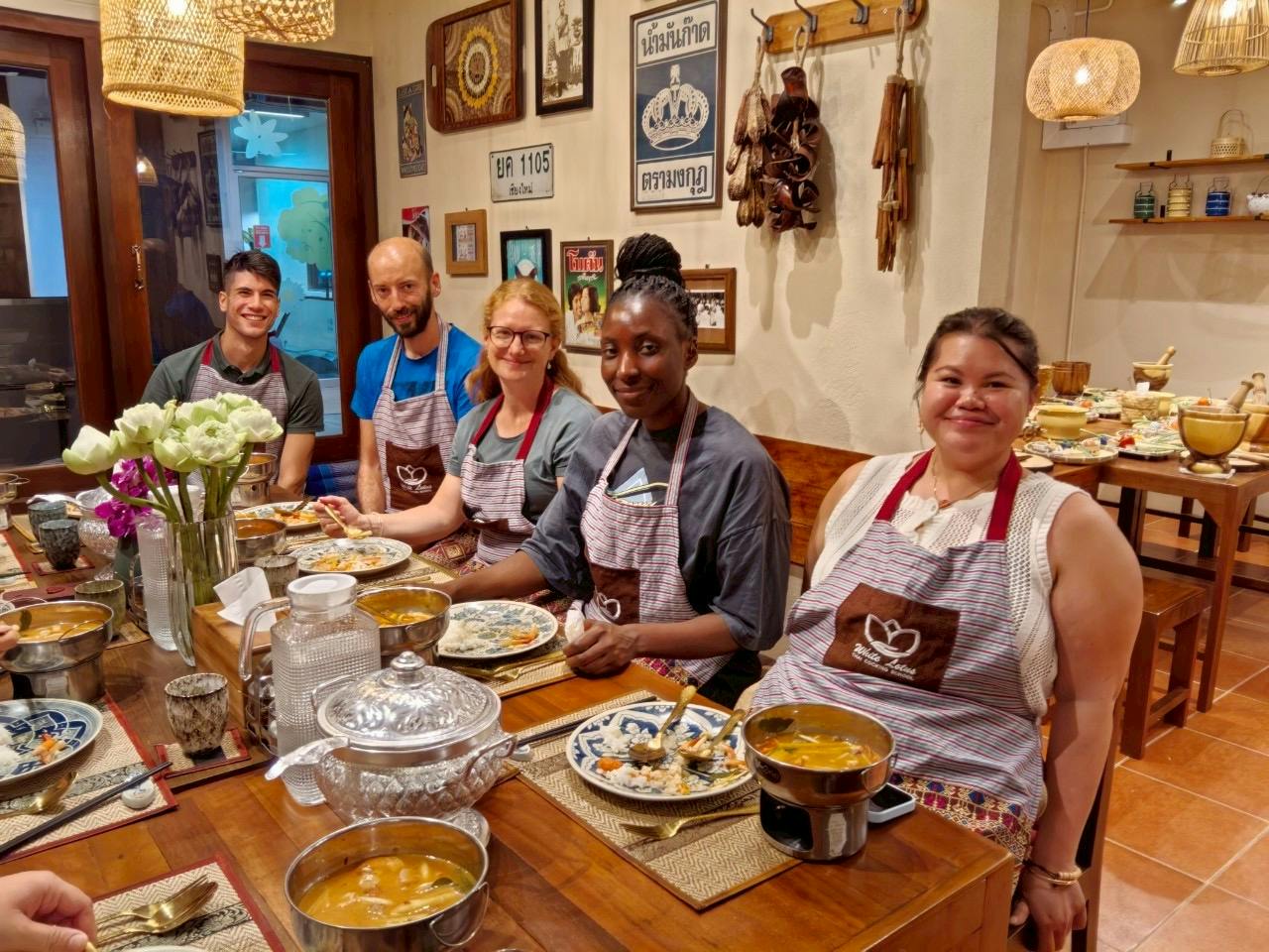 Four people wearing aprons sit at a dining table with plates of food and dishes in a cozy, decorated room.