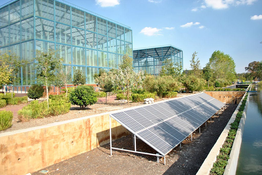 Pannelli solari a terra vicino a moderni edifici in vetro circondati da alberi e da un canale d'acqua sotto un cielo azzurro con nuvole.