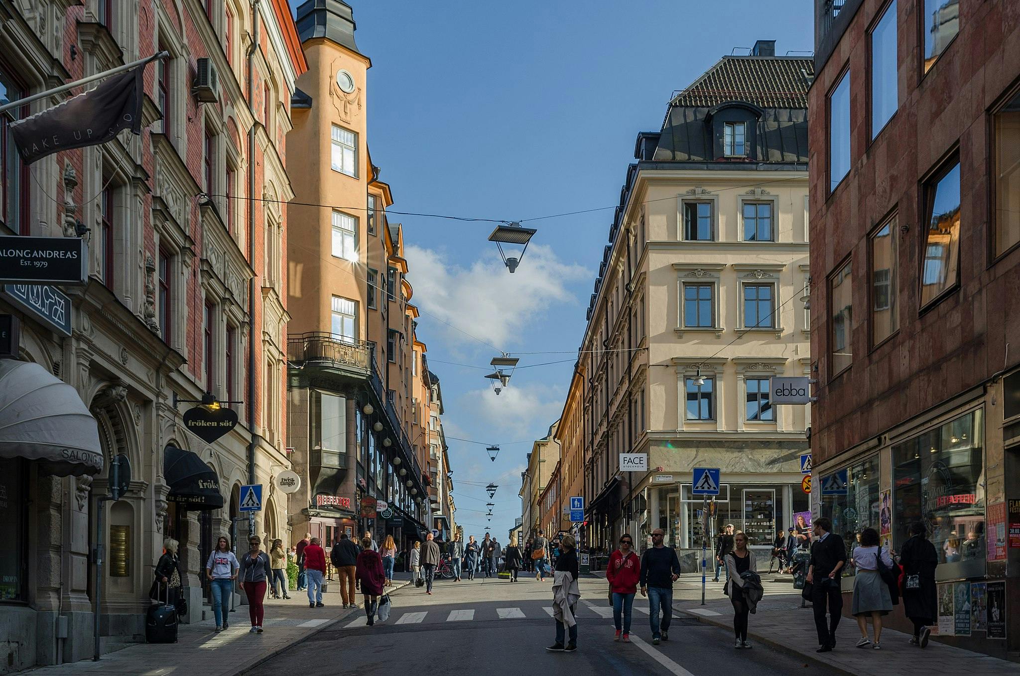 맑고 푸른 하늘 아래 높은 건물과 상점이 늘어선 좁은 골목길을 걷는 사람들.
