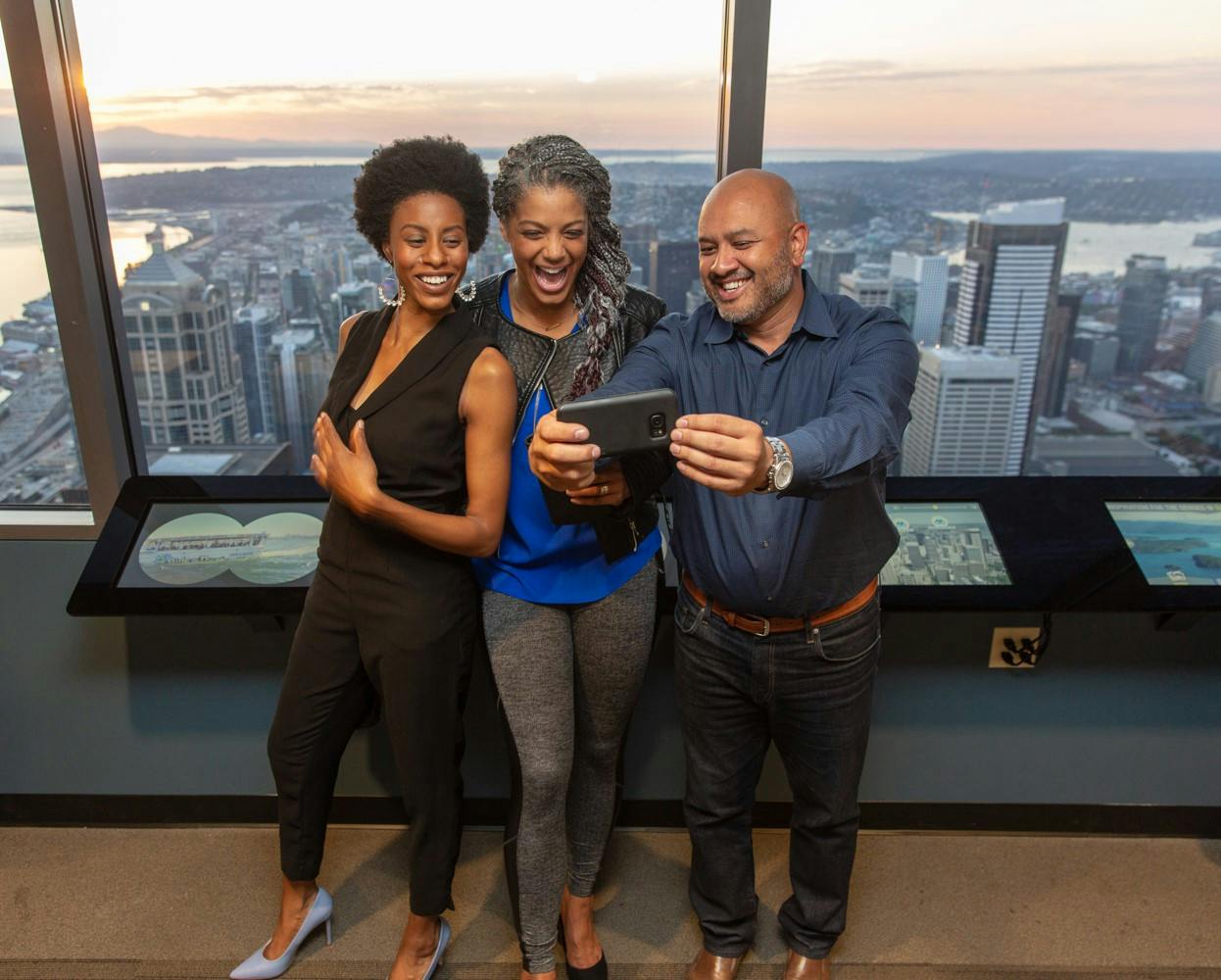 Три человека улыбаются и делают селфи на фоне городского пейзажа на закате, если смотреть с крытой смотровой площадки.