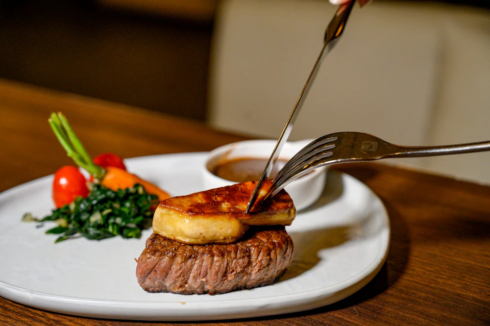 A plate with a steak topped with seared foie gras, served with spinach, vegetables, and sauce in a white dish.