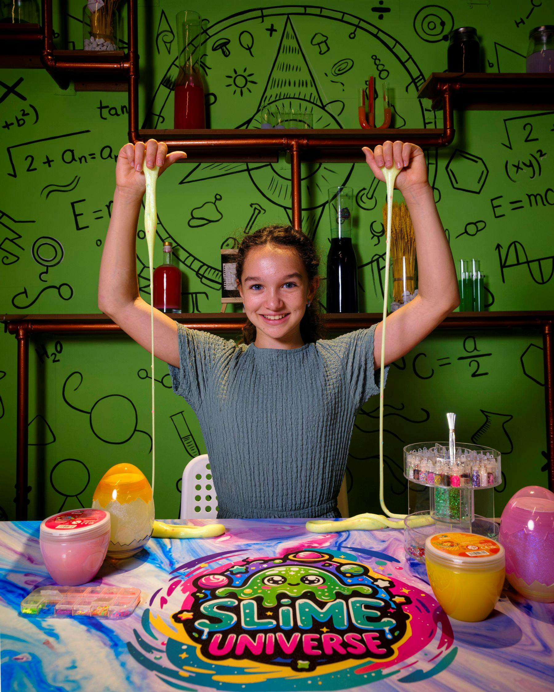 Ένα άτομο με πράσινο πουκάμισο που κρατά τεντωμένη γλίτσα, καθισμένο σε ένα πολύχρωμο τραπέζι με διάφορα δοχεία, σε ένα δωμάτιο με θέμα την επιστήμη.