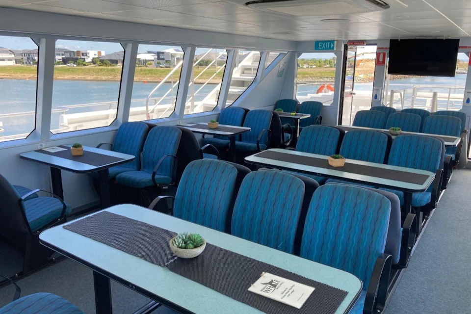Passenger seating area on a boat with blue chairs and tables, small potted plants, and large windows showing a waterfront view.