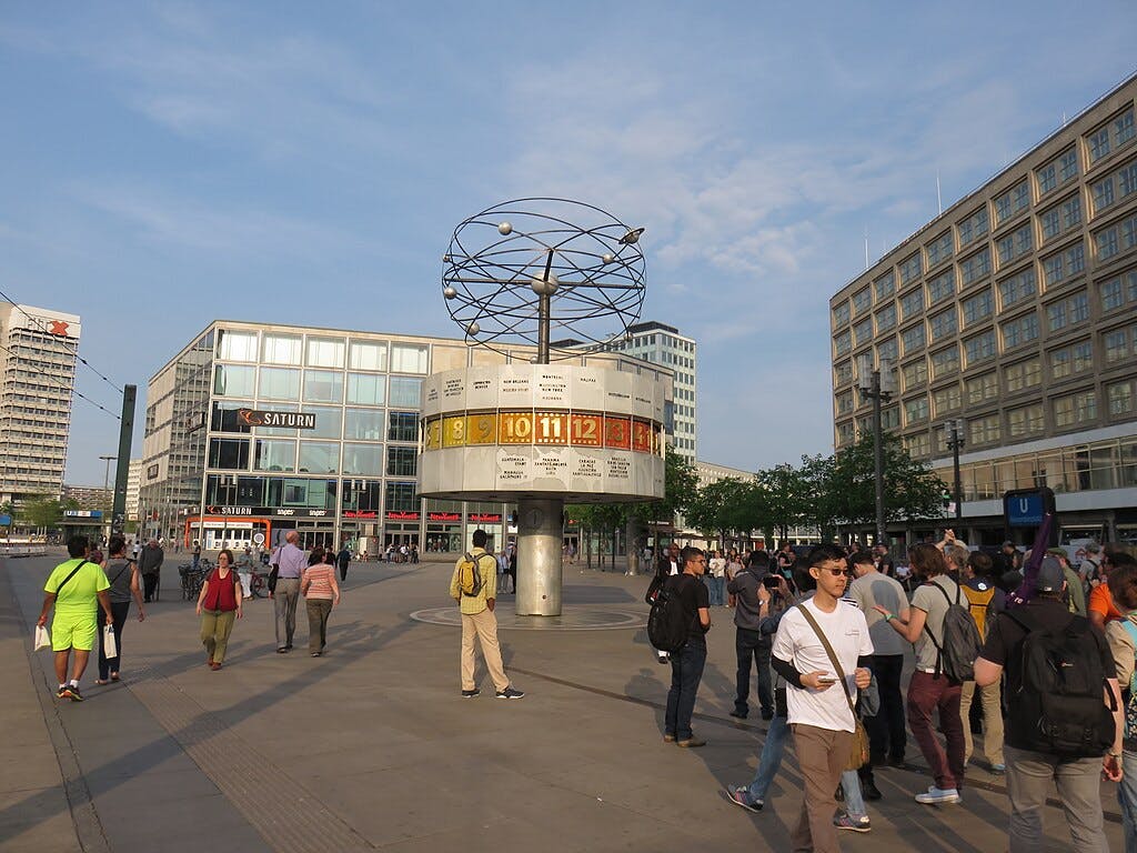 Crowded urban plaza with people walking and gathering, featuring a large world clock structure surrounded by modern buildings.