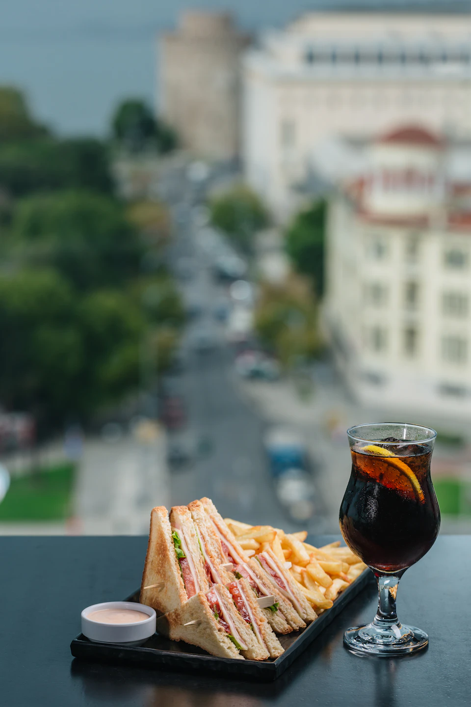 Club sandwich, fries, and a glass of soda with lemon on a table, overlooking a city street and buildings in the background.