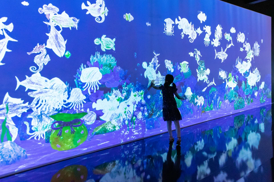 A person in a dark dress interacts with a large digital underwater display featuring colorful marine life and plants.