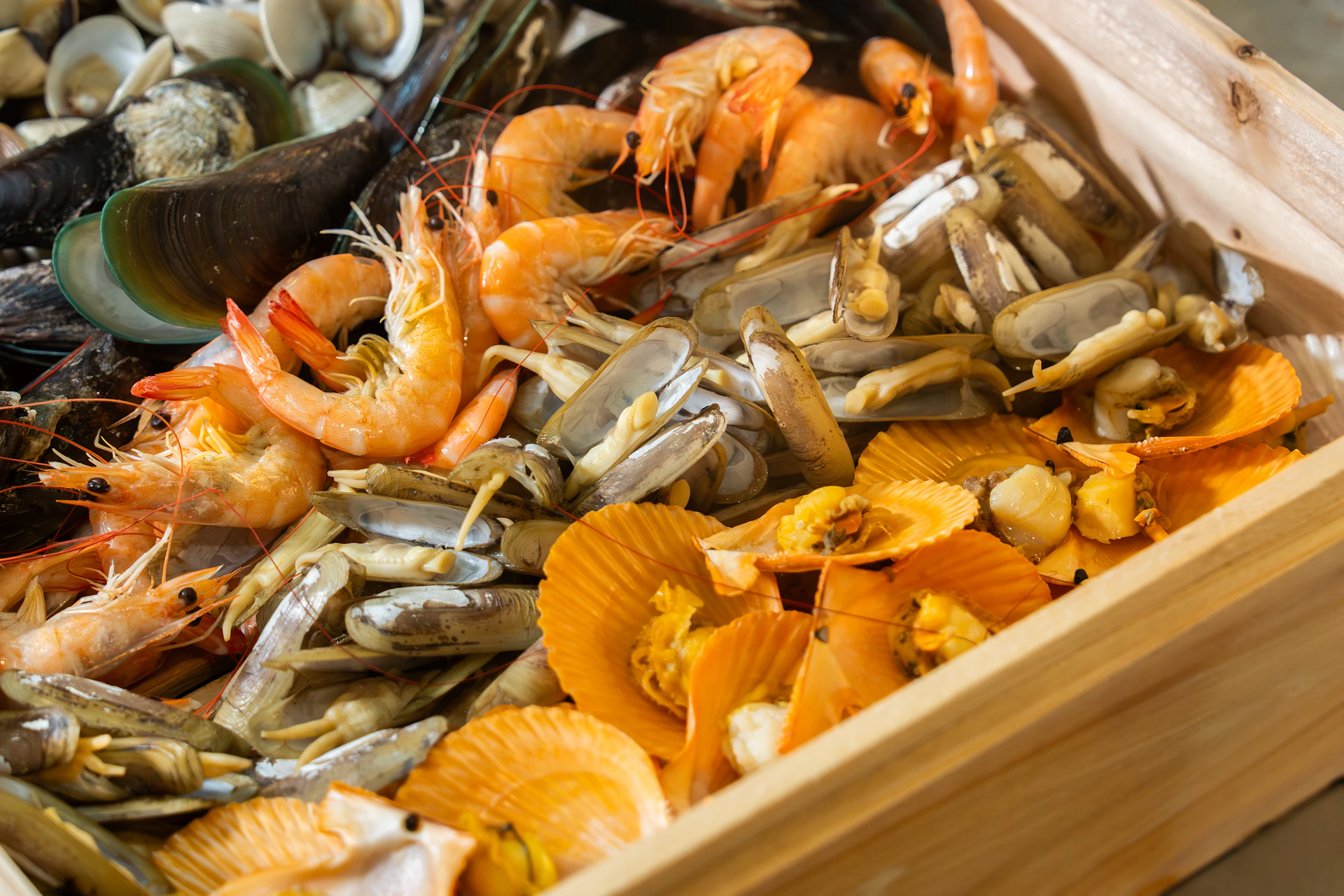 A wooden tray filled with seafood, including shrimp, clams, mussels, and scallops in shells.