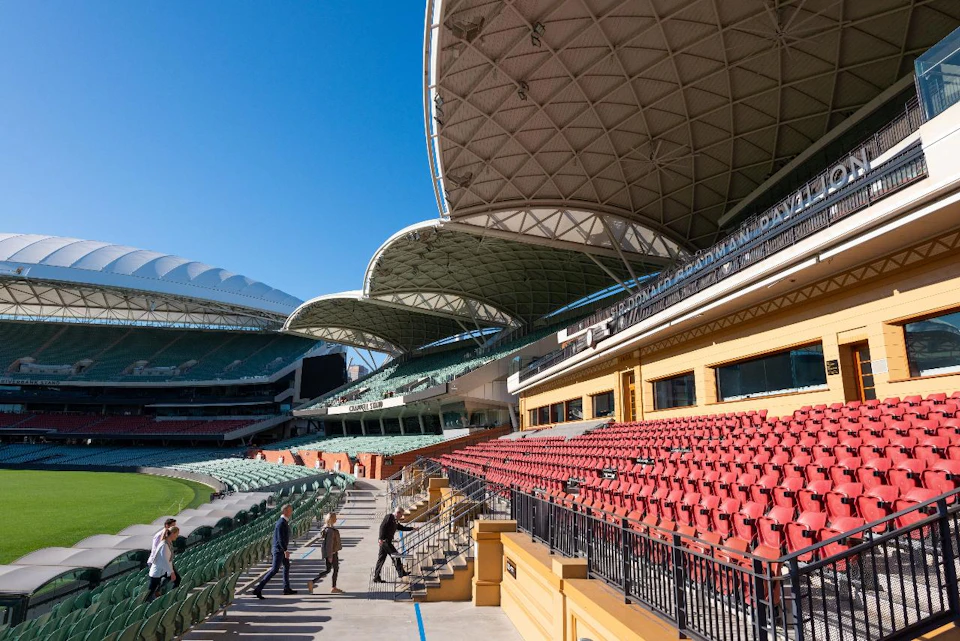 Modern stadium with distinctive curved roof, empty red and green seats, and a few people walking near the field. Clear blue sky overhead.