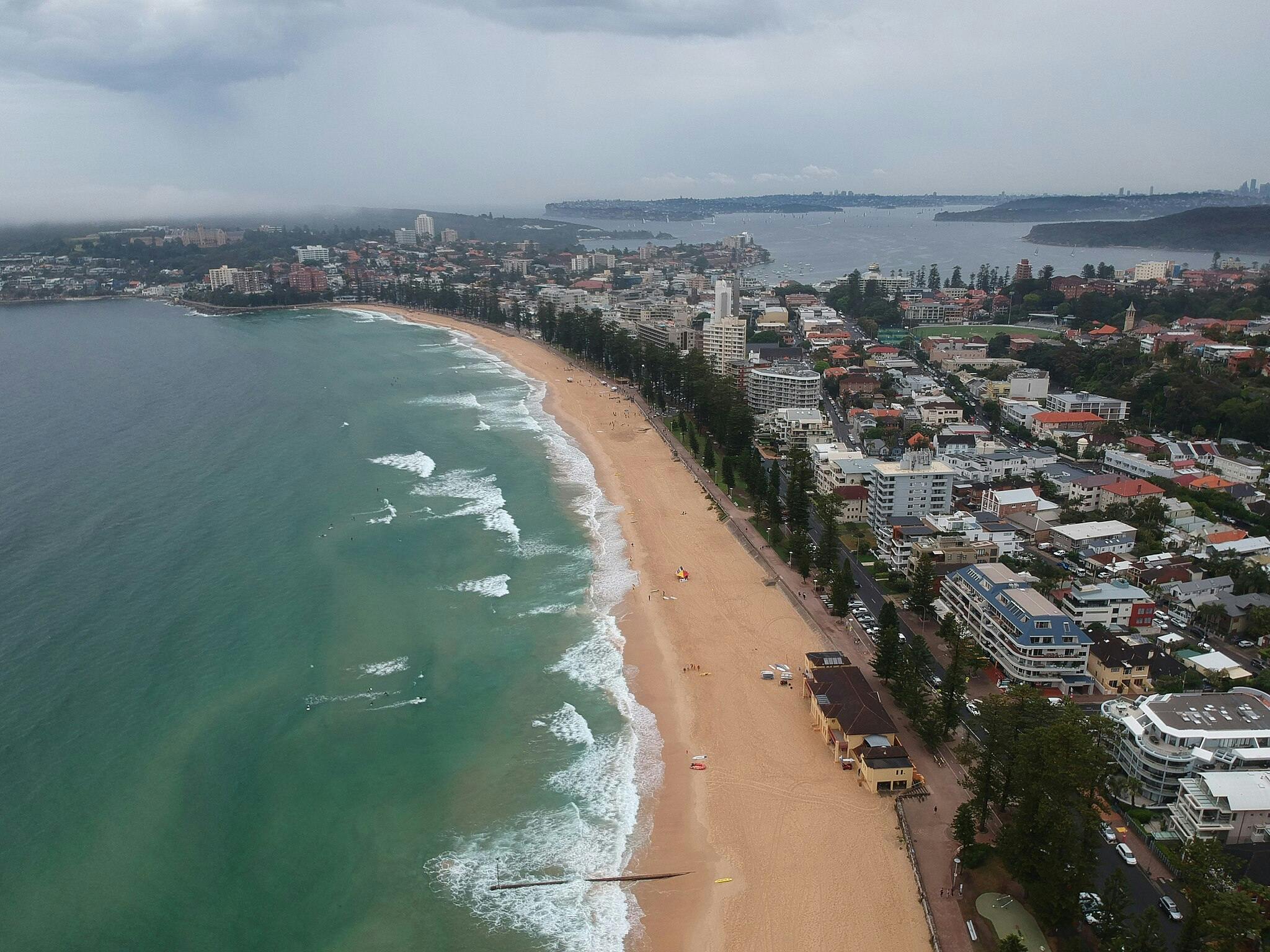 Widok z lotu ptaka na nadmorskie miasto z piaszczystą plażą z jednej strony i budynkami miejskimi z drugiej, graniczącymi z oceanem.