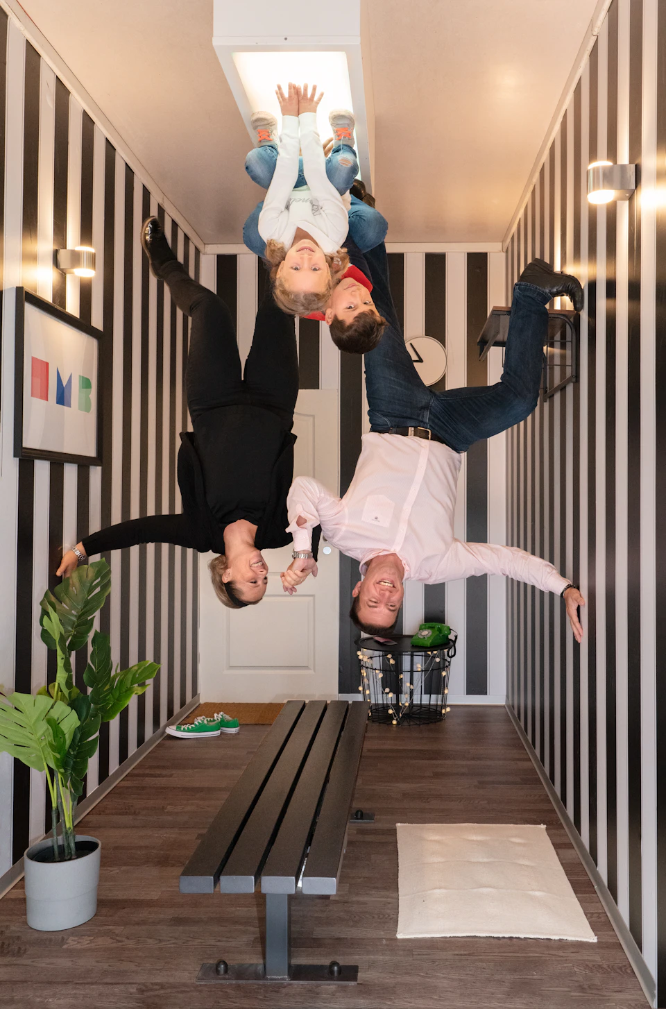 Four people appear to hang upside down in a room with black and white striped walls, creating an optical illusion.