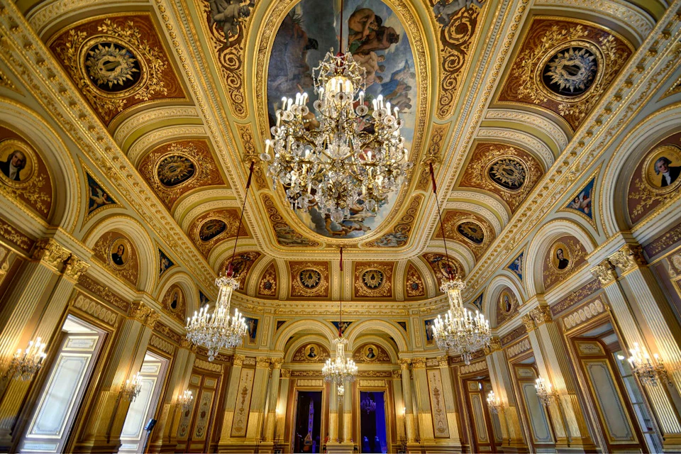 A lavish, ornately decorated room with chandeliers, arched windows, and a painted ceiling in a classical style.