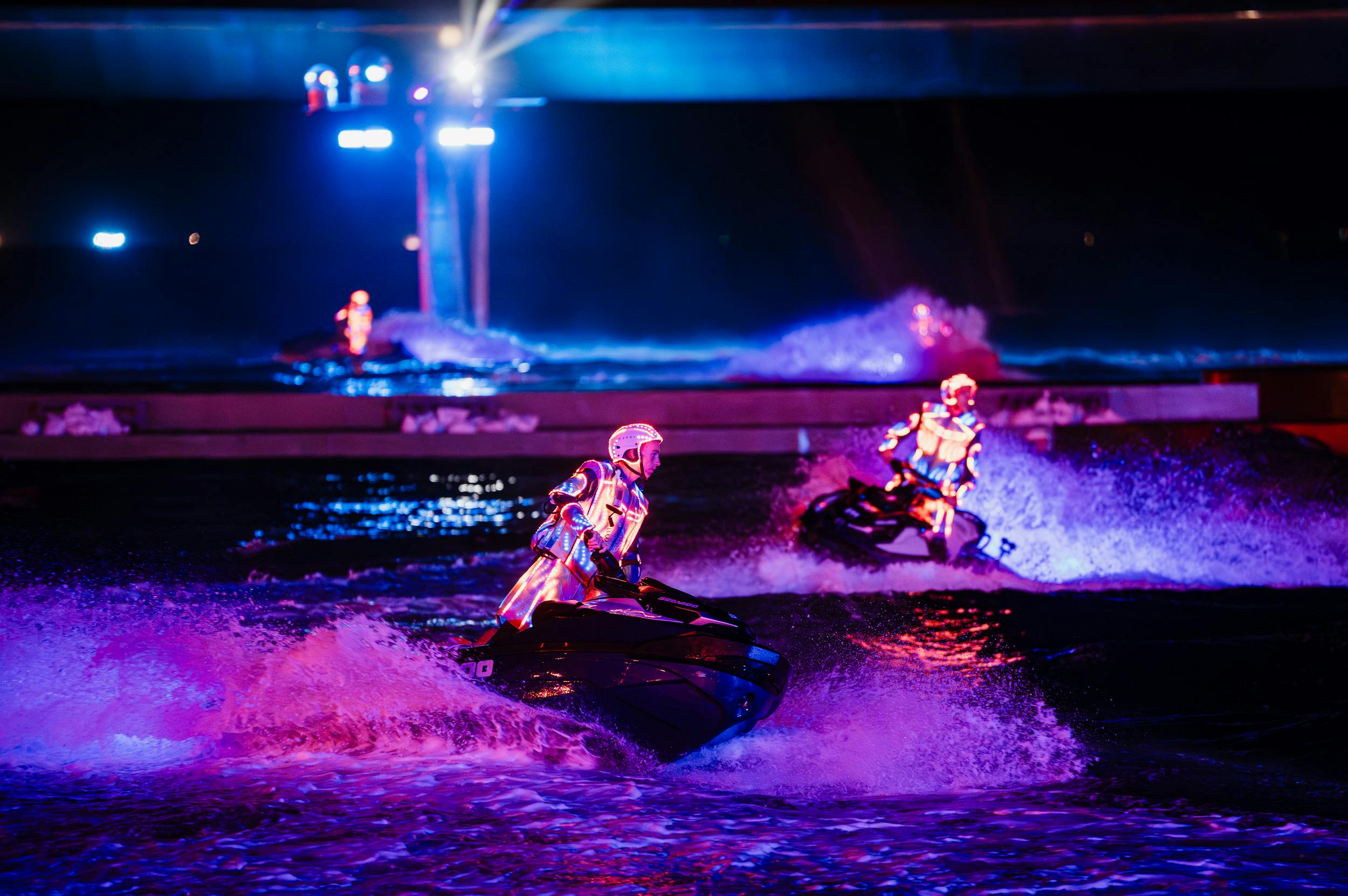 Performers in illuminated suits riding jet skis on water at night under colorful lights near a bridge.