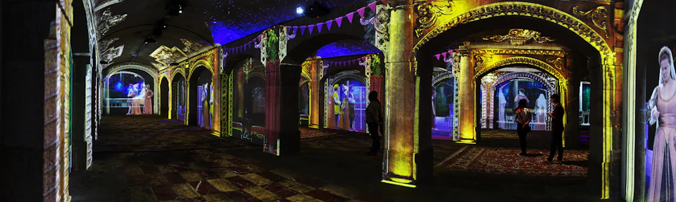 A person stands in a dark room with illuminated archways and columns, decorated with projections of people and medieval scenes.