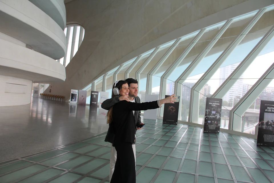 Two people inside a modern building with large windows, taking a selfie. One is wearing headphones. Display panels are in the background.