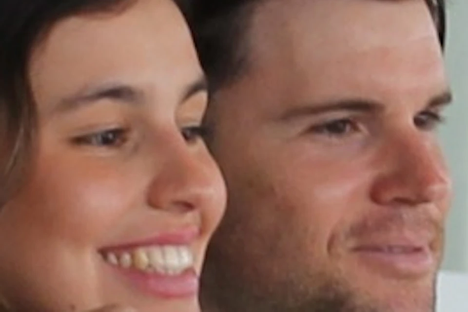 Two people inside a modern building with large windows, taking a selfie. One is wearing headphones. Display panels are in the background.