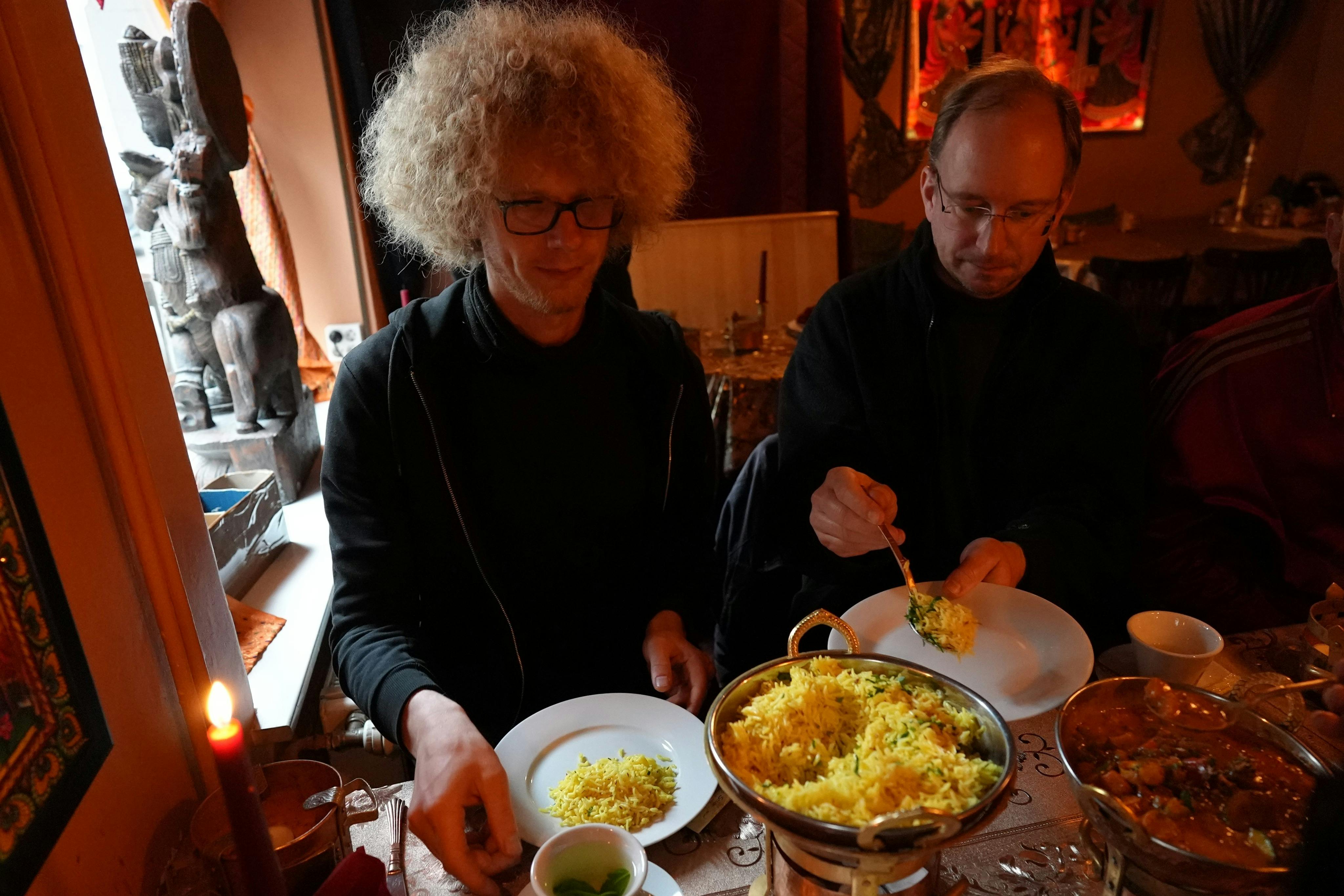 To mænd sidder ved et bord og serverer sig selv fra fælles retter med gule ris og karry.