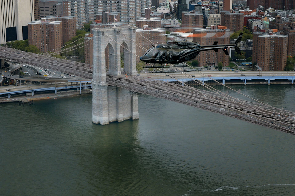 A helicopter flies over the Brooklyn Bridge with the cityscape and river in the background.