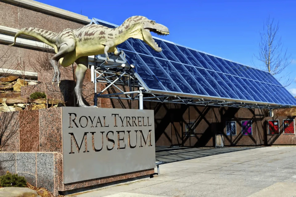 A dinosaur statue above the "Royal Tyrrell Museum" sign, with rows of large glass panels on the adjacent building.