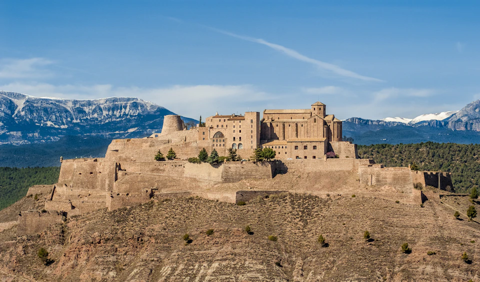 A large, tan-colored historic fortress on a hilltop with mountains and a blue sky in the background.