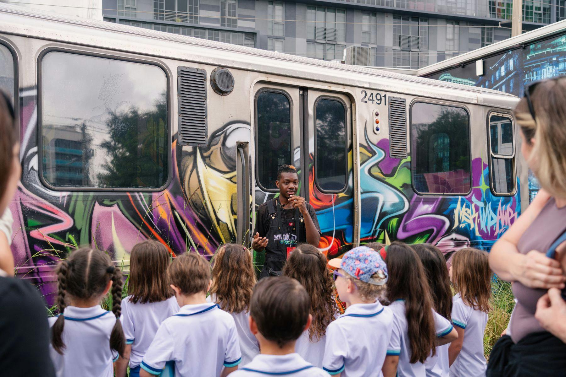 一名男子在地铁列车前与一群孩子交谈，车上涂满了五颜六色的涂鸦。