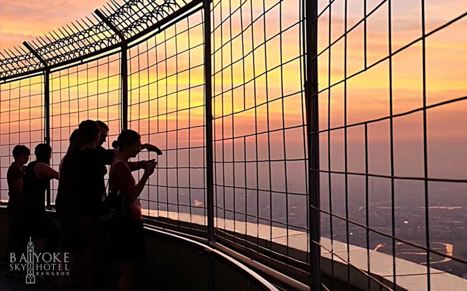 Two people stand near a fenced observation deck, one holding a phone, against a vibrant sunset sky.