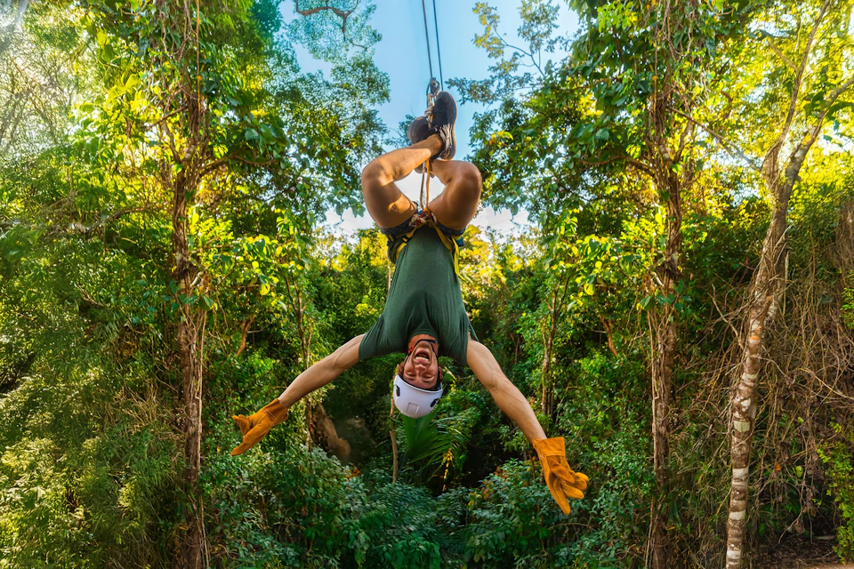 Person hanging upside down on a zipline in a forested area, wearing a helmet, gloves, and a harness, smiling.