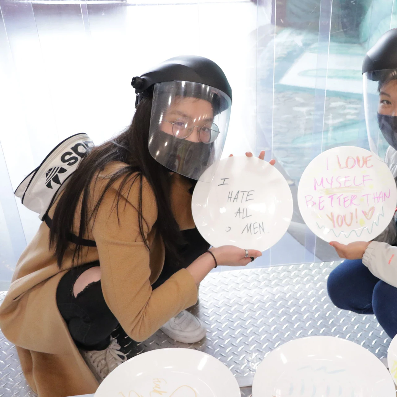 People wearing face shields, holding plates with handwritten messages: "I HATE ALL MEN" and "I LOVE MYSELF BETTER THAN YOU!"