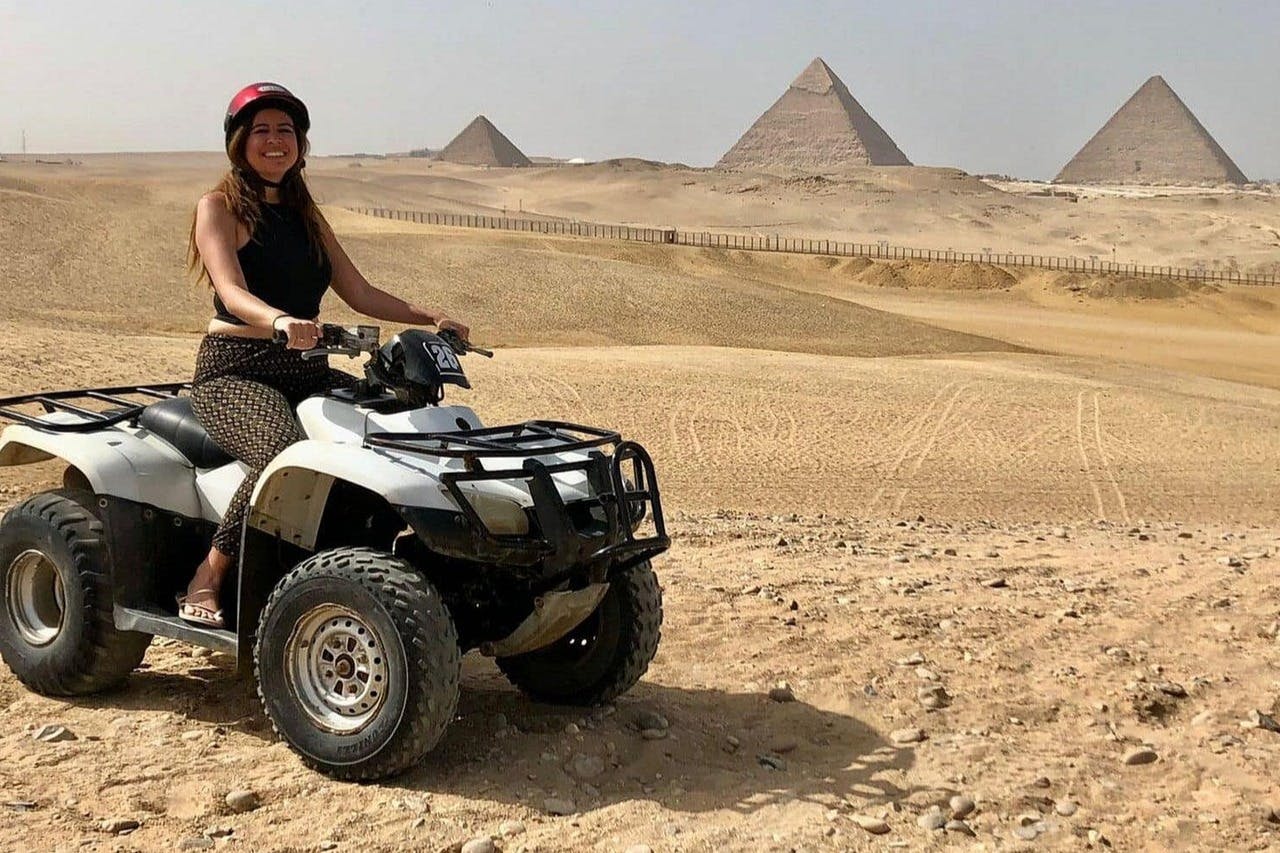 赤いヘルメットをかぶった女性が砂漠でクワッドバイクに乗っています。背景にはいくつかのピラミッドが見えます。
