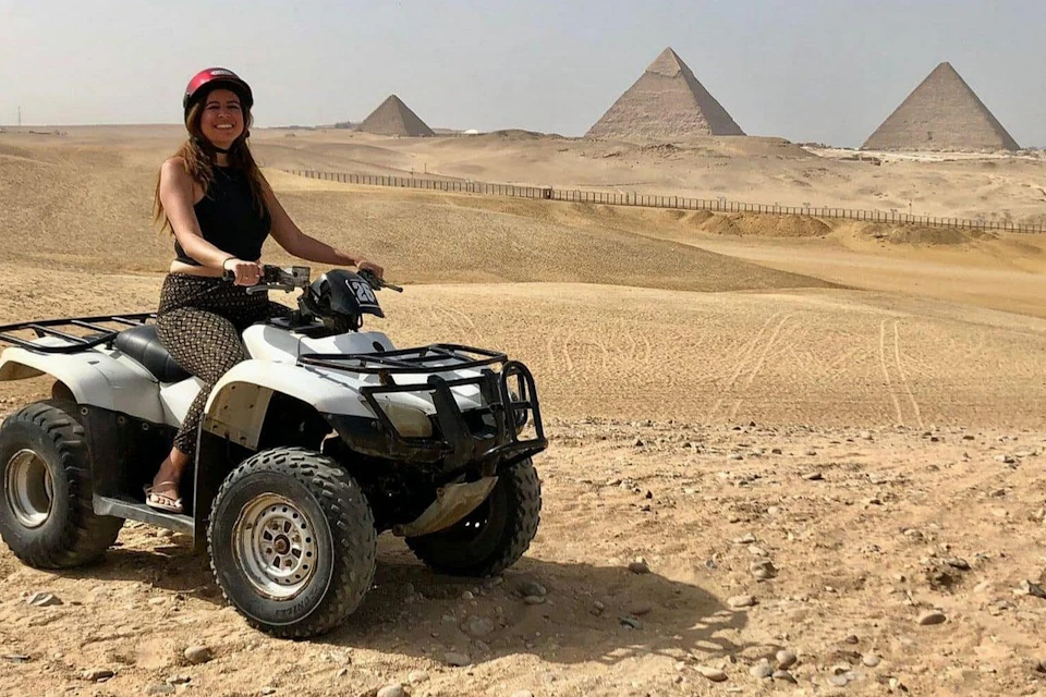 A woman in a red helmet rides a quad bike in a desert with several pyramids visible in the background.