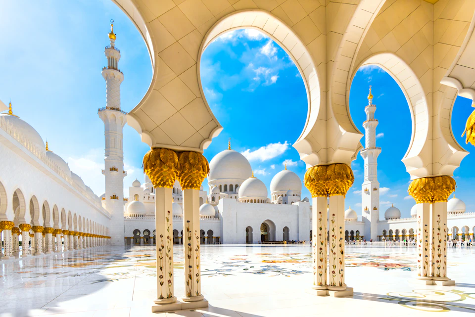 Intricate white and gold arches frame a courtyard with domes and minarets under a bright blue sky with some clouds.