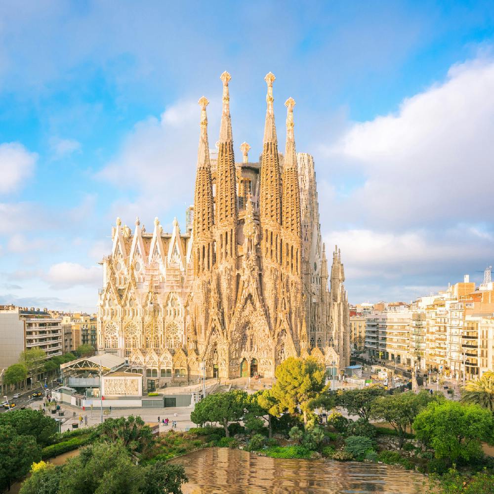 Vue aérienne de la basilique de la Sagrada Família à Barcelone avec le paysage urbain environnant, la verdure et un étang au premier plan.