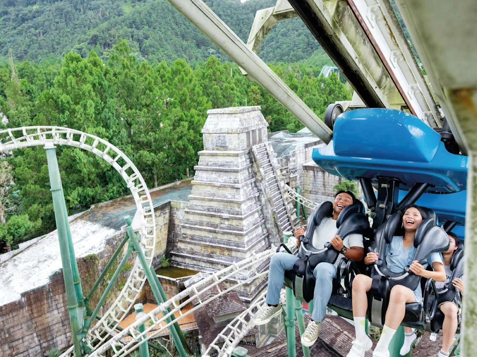 People on a blue rollercoaster ride, smiling and enjoying the experience, with greenery and structures in the background.