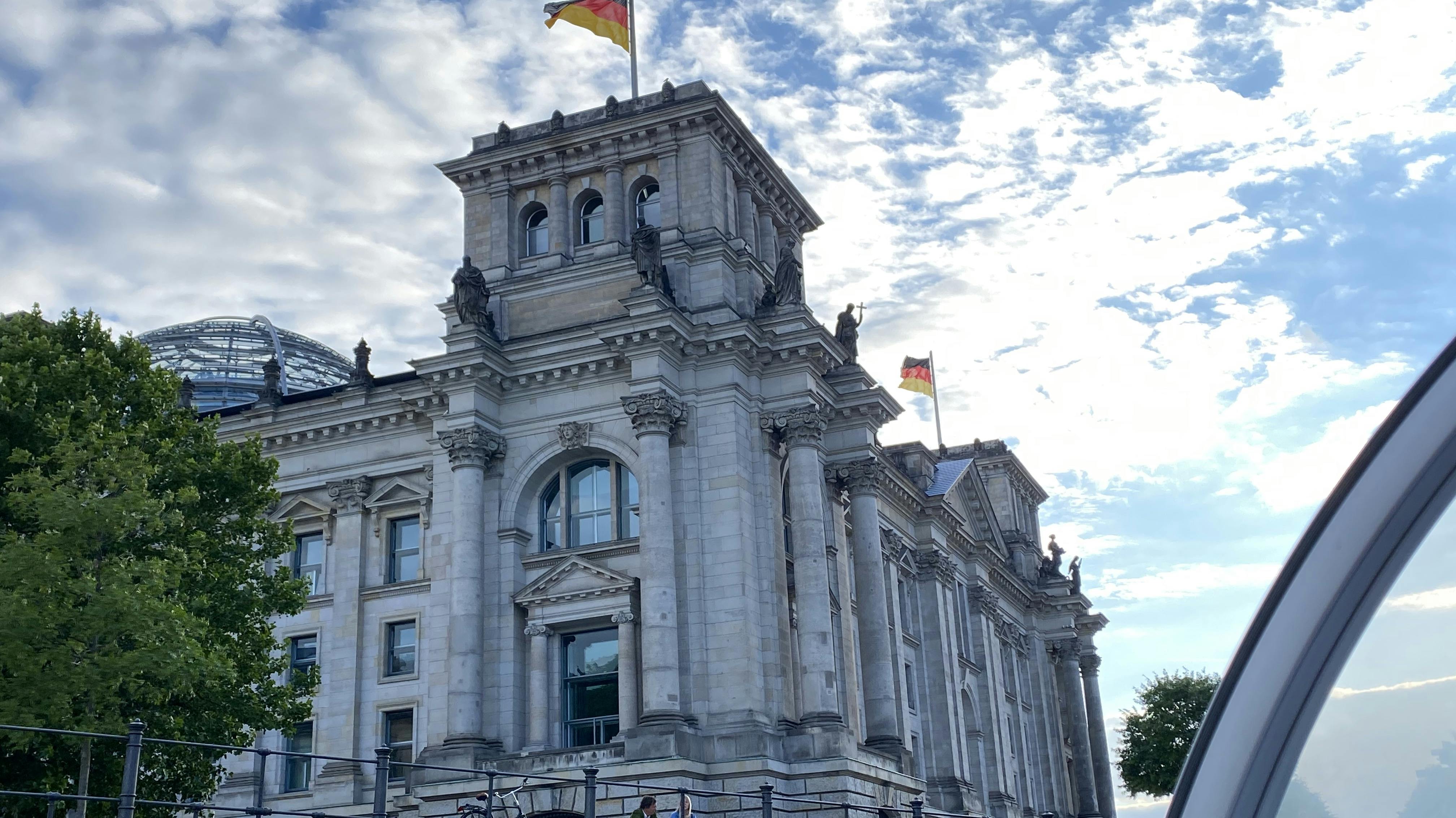 Ιστορικό κτίριο με κίονες και περίτεχνες λεπτομέρειες, δύο σημαίες στην κορυφή, κάτω από έναν εν μέρει συννεφιασμένο ουρανό.