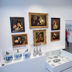 Ett galleri med inramade målningar, keramiska vaser och små artefakter i montrar; en person går i korridoren.