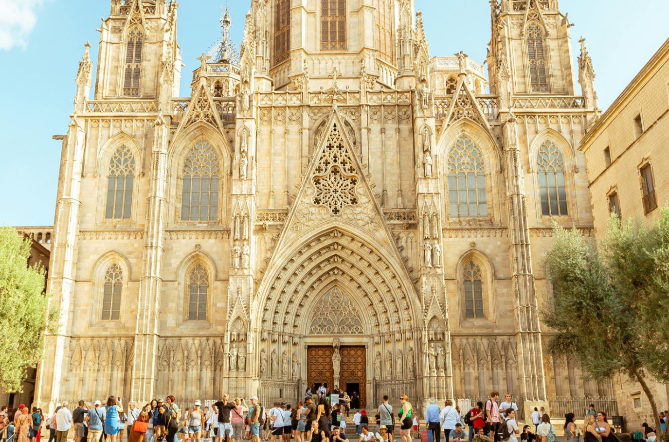 La facciata di una grande cattedrale gotica con dettagli intricati, ingressi ad arco e numerosi turisti radunati di fronte in una giornata di sole.