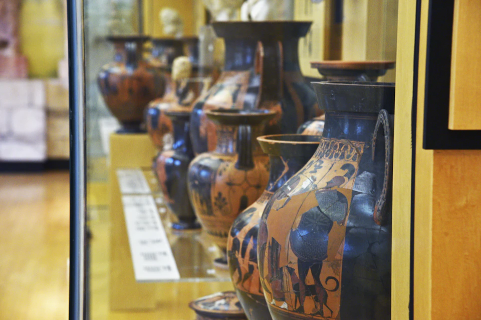 A display of ancient, painted pottery vases in a museum setting, arranged on shelves with informational plaques.