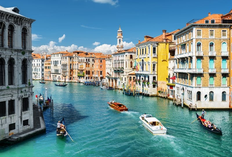 Canal vénitien avec gondoles et bateaux à moteur, bordé de bâtiments historiques colorés sous un ciel bleu clair.