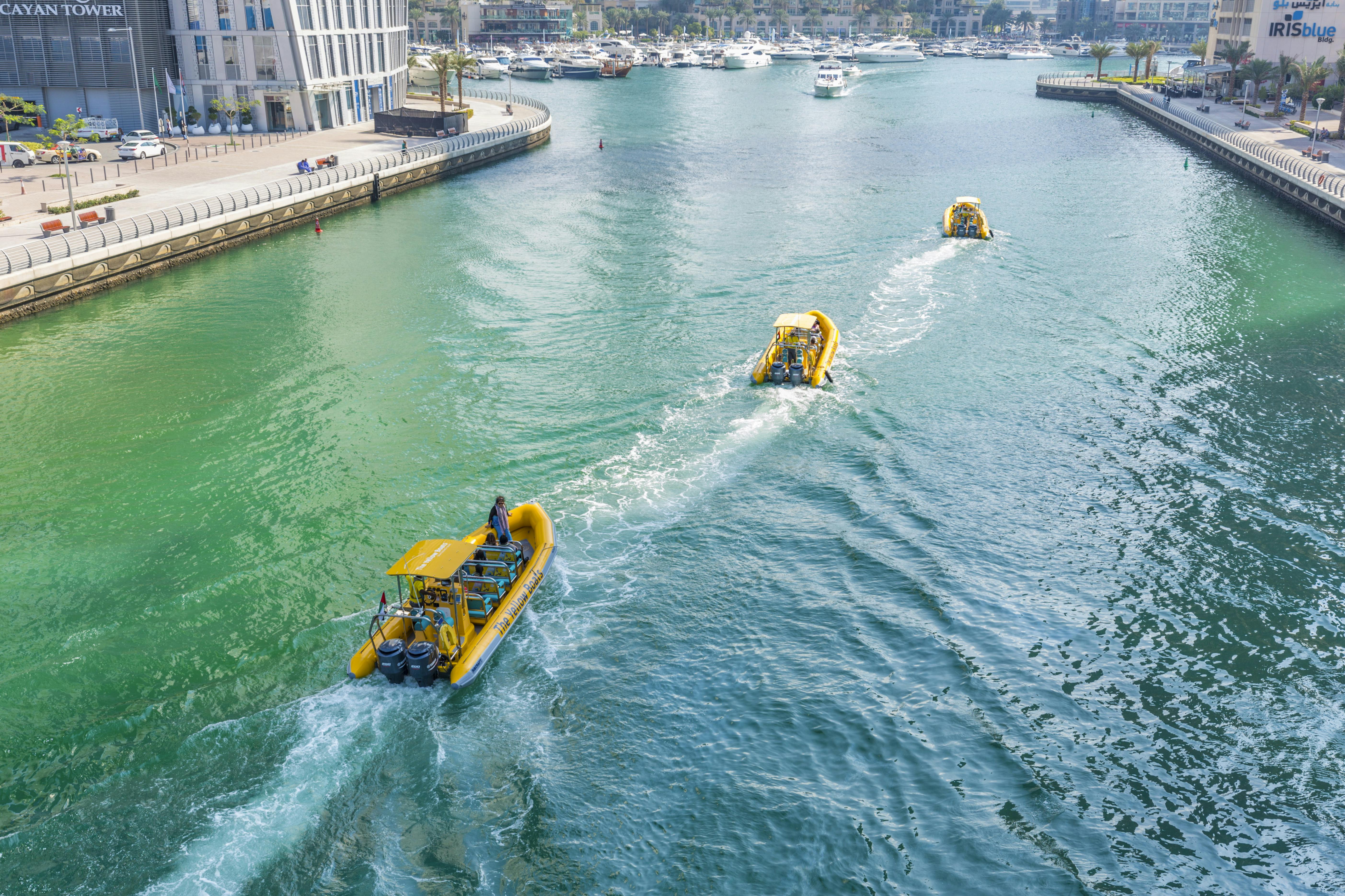 Tři žluté motorové čluny na zelenomodrém vodním kanálu s budovou na nábřeží a četnými zakotvenými čluny v pozadí.