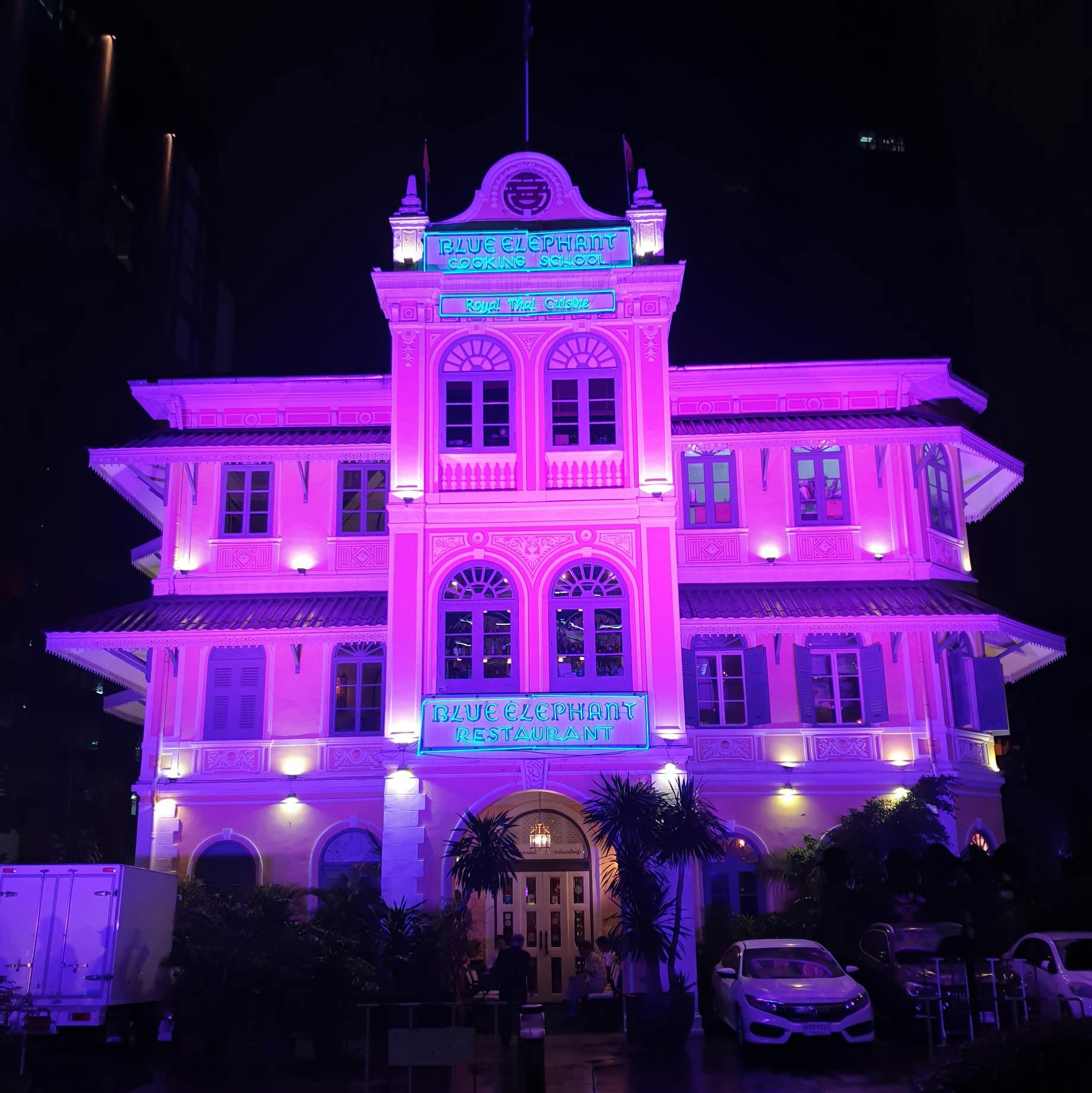 Un bâtiment de style colonial très éclairé avec des enseignes au néon pour "Blue Elephant Cooking School" et "Blue Elephant Restaurant" la nuit.